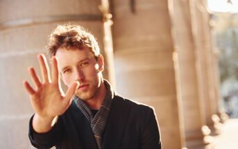 A young man with curly hair and stubble extends his hand toward the camera as if to stop or block the view. He wears a dark blazer and a scarf while standing in front of large stone columns bathed in warm sunlight. The background is out of focus.