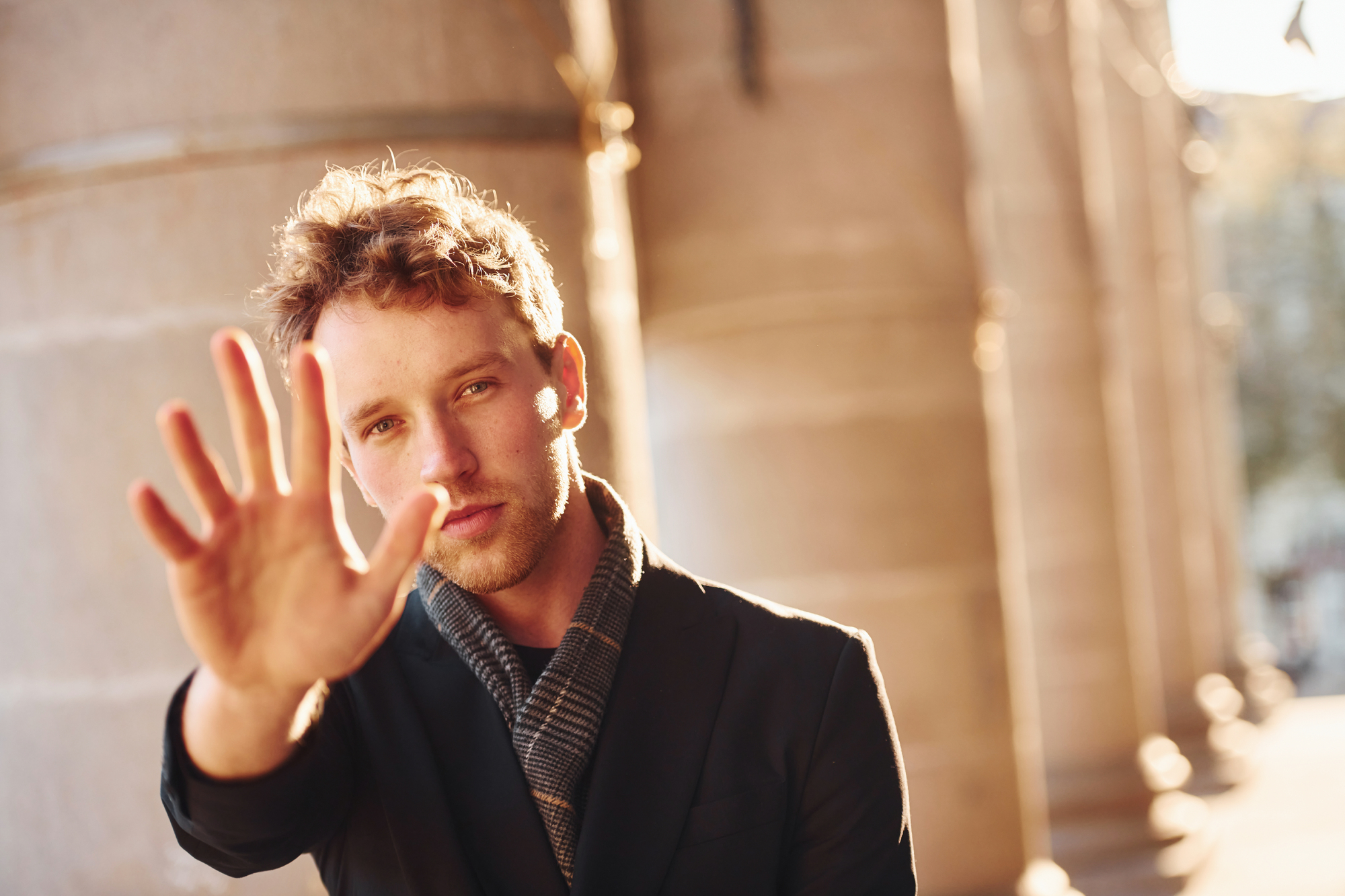 A young man with curly hair and stubble extends his hand toward the camera as if to stop or block the view. He wears a dark blazer and a scarf while standing in front of large stone columns bathed in warm sunlight. The background is out of focus.