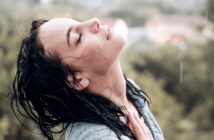 A woman with wet hair tilts her head back and closes her eyes, enjoying the sensation of raindrops on her face. She is outside, with a blurred green and brown background indicating nature. Her hand gently touches her chest, and droplets of water are visible on her face.