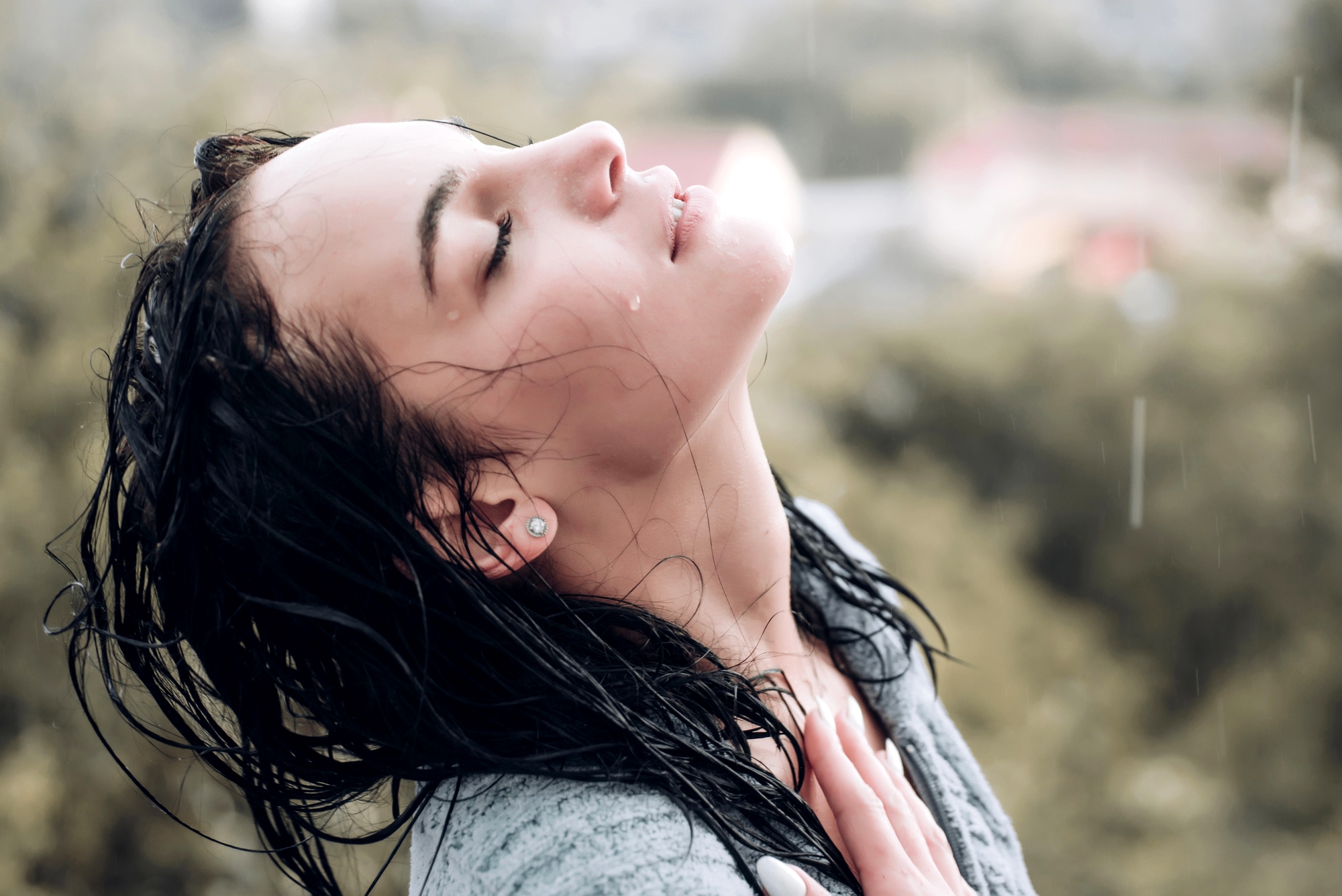 A woman with wet hair tilts her head back and closes her eyes, enjoying the sensation of raindrops on her face. She is outside, with a blurred green and brown background indicating nature. Her hand gently touches her chest, and droplets of water are visible on her face.