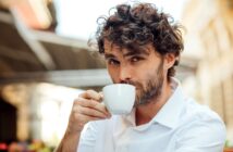 A man with curly brown hair and a beard, wearing a white shirt, sits outdoors and sips from a white coffee cup. He looks directly into the camera, and the blurred background suggests a street or café setting.