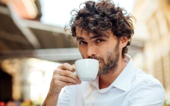 A man with curly brown hair and a beard, wearing a white shirt, sits outdoors and sips from a white coffee cup. He looks directly into the camera, and the blurred background suggests a street or café setting.