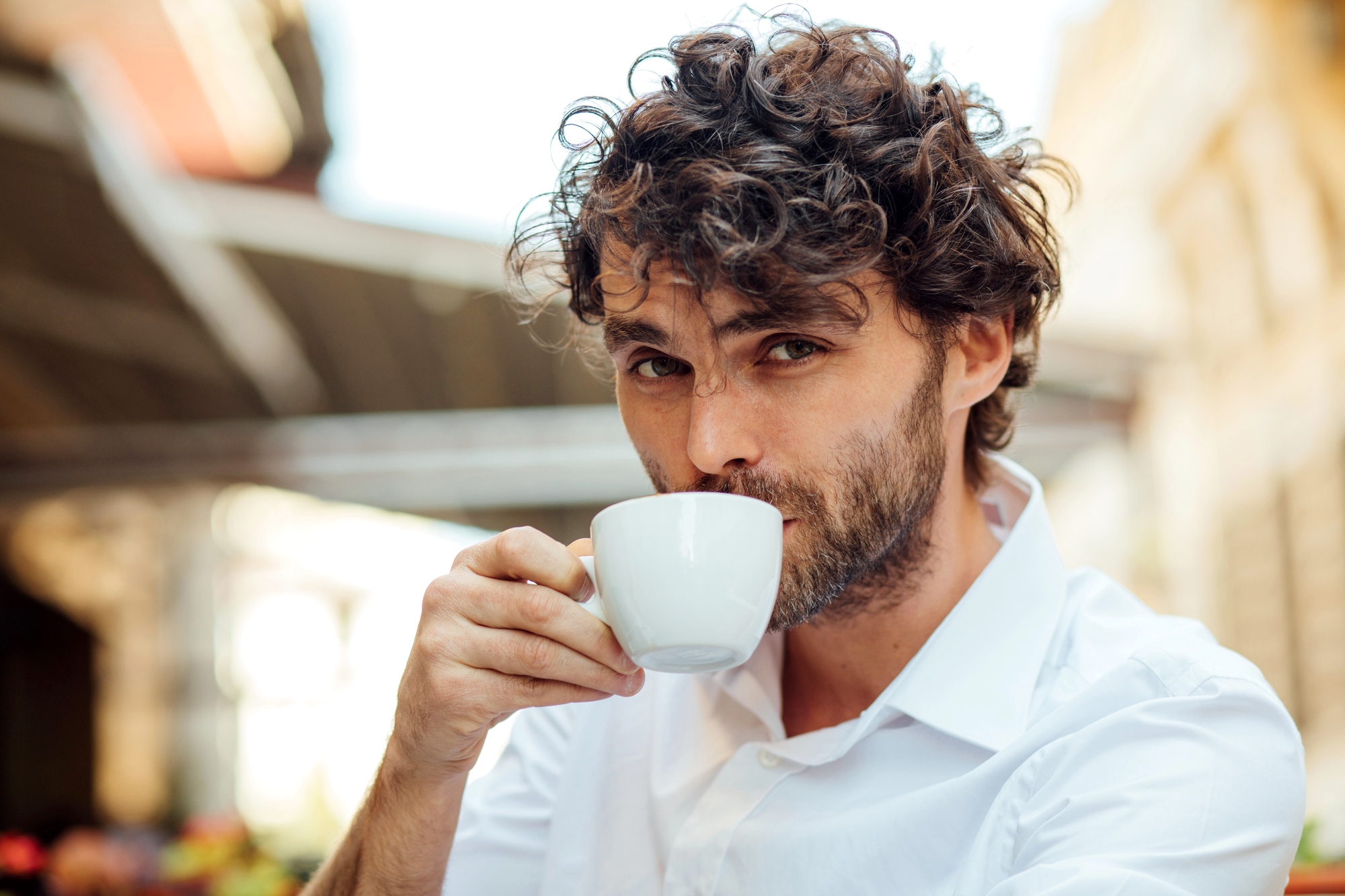 A man with curly brown hair and a beard, wearing a white shirt, sits outdoors and sips from a white coffee cup. He looks directly into the camera, and the blurred background suggests a street or café setting.