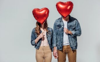 Two people standing side by side, each holding a red heart-shaped balloon in front of their faces, obscuring their identities. They are holding hands and wearing matching outfits: denim jackets with beige pants. The background is plain white.