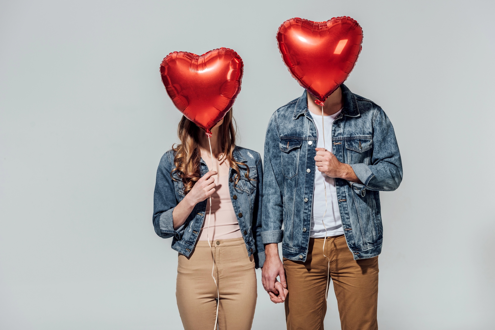 Two people standing side by side, each holding a red heart-shaped balloon in front of their faces, obscuring their identities. They are holding hands and wearing matching outfits: denim jackets with beige pants. The background is plain white.