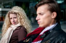 A woman with curly blonde hair and a serious expression looks at a man with straight brown hair who is out of focus in the foreground. Both are dressed warmly, suggesting a cool day. The background appears to be an urban setting with modern buildings.