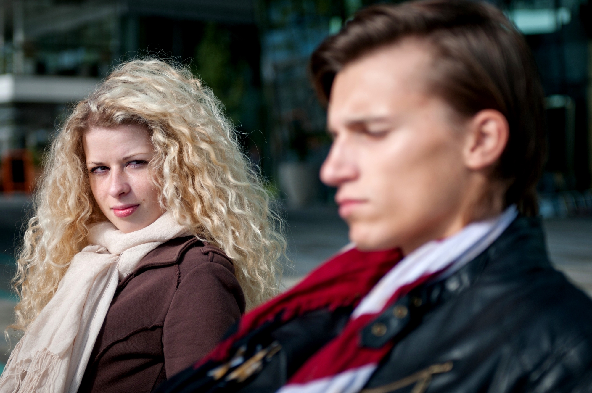 A woman with curly blonde hair and a serious expression looks at a man with straight brown hair who is out of focus in the foreground. Both are dressed warmly, suggesting a cool day. The background appears to be an urban setting with modern buildings.