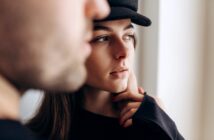 Close-up of a contemplative young woman wearing a black hat, partially obscured by a blurry figure in the foreground. She's looking to the right, with her hand gently touching her face. The background is softly lit.