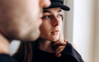 Close-up of a contemplative young woman wearing a black hat, partially obscured by a blurry figure in the foreground. She's looking to the right, with her hand gently touching her face. The background is softly lit.