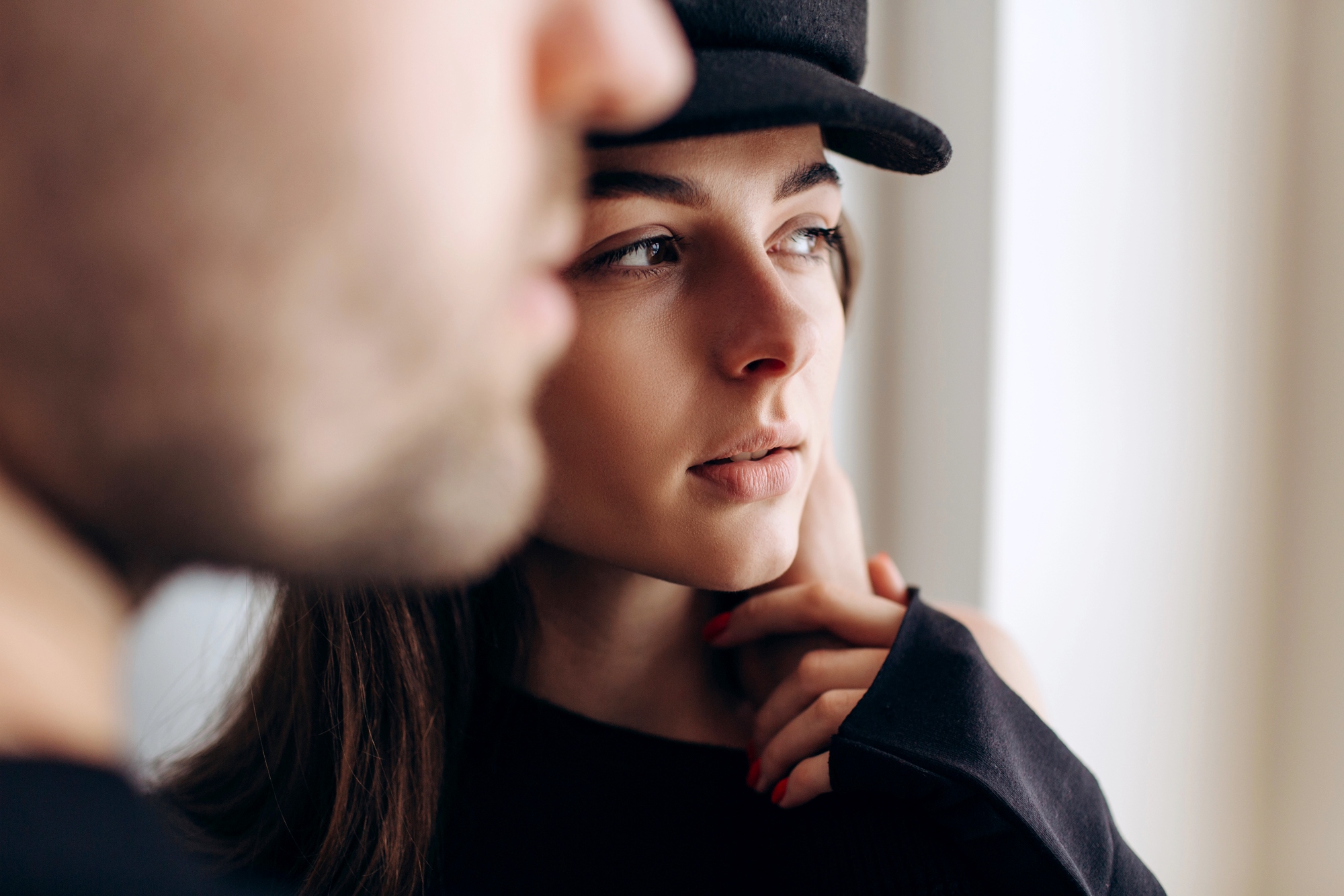 Close-up of a contemplative young woman wearing a black hat, partially obscured by a blurry figure in the foreground. She's looking to the right, with her hand gently touching her face. The background is softly lit.