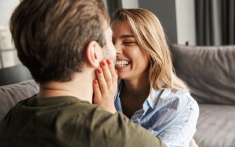 A woman with long blonde hair and a striped shirt smiles while gently touching the face of a man with brown hair and a green shirt. They are sitting close together on a sofa, sharing an affectionate moment.