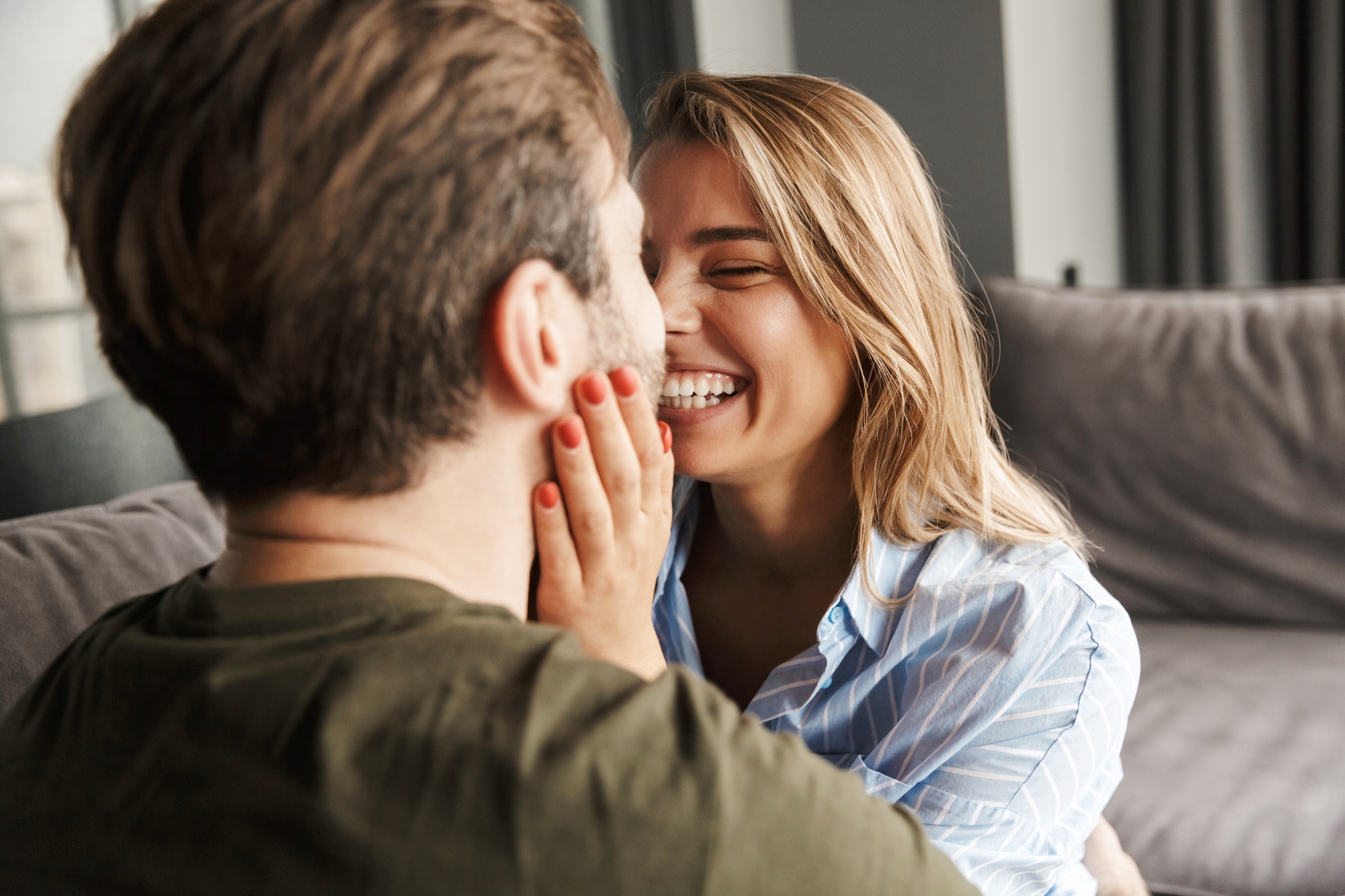 A woman with long blonde hair and a striped shirt smiles while gently touching the face of a man with brown hair and a green shirt. They are sitting close together on a sofa, sharing an affectionate moment.