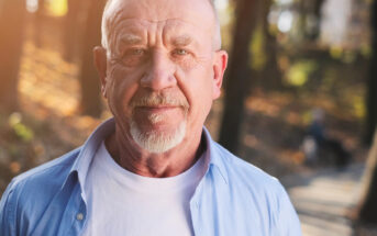 A bald elderly man with a white beard and mustache is wearing a white T-shirt and an unbuttoned light blue shirt. He stands in a sunlit park with a mix of sunlight and shadows falling on him, and trees and a path visible in the background.