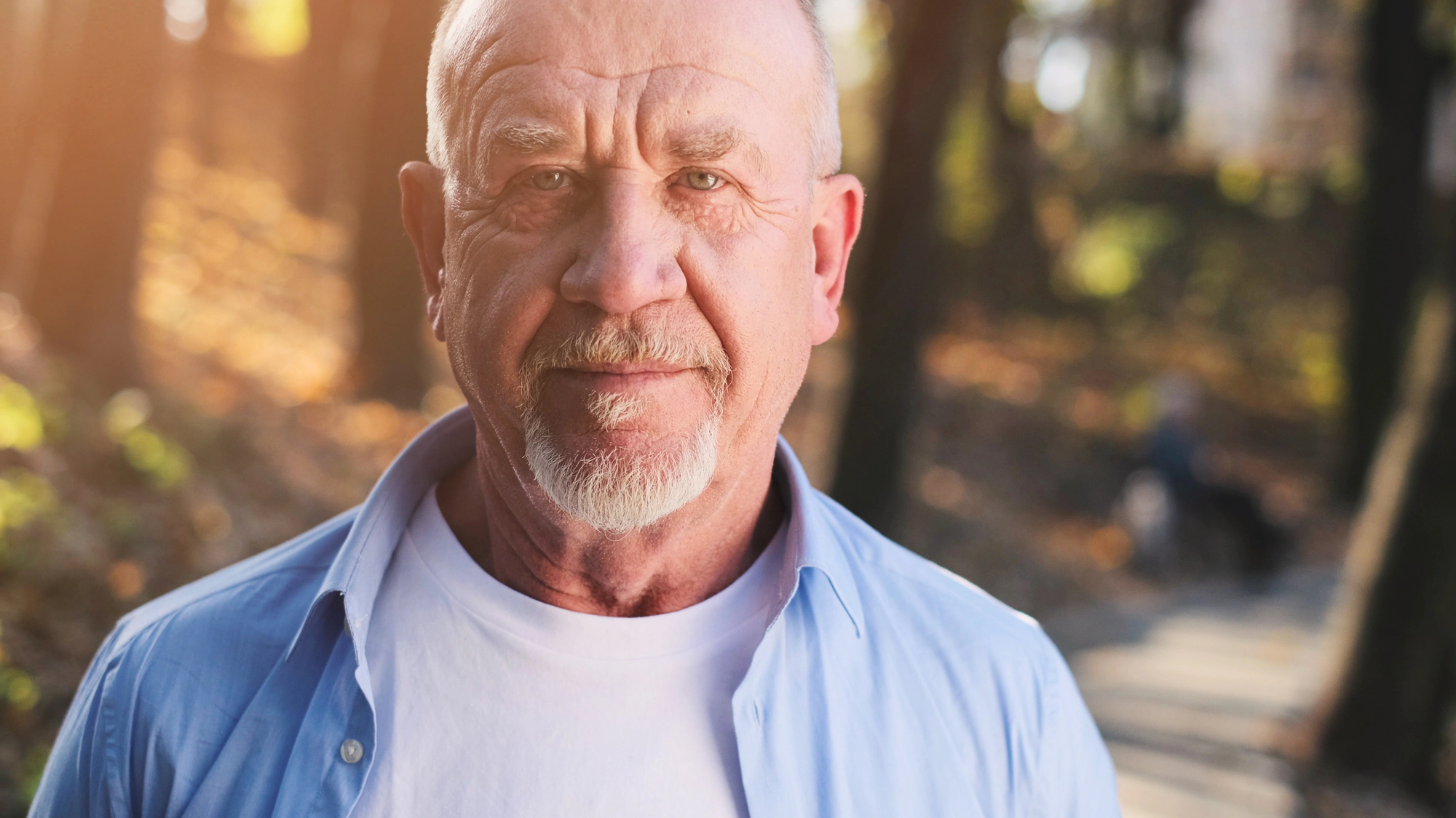 A bald elderly man with a white beard and mustache is wearing a white T-shirt and an unbuttoned light blue shirt. He stands in a sunlit park with a mix of sunlight and shadows falling on him, and trees and a path visible in the background.