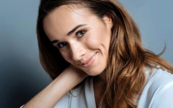 A woman with long brown hair and a light complexion smiles softly while resting her head on her hand. She is wearing a white shirt and has a neutral background.