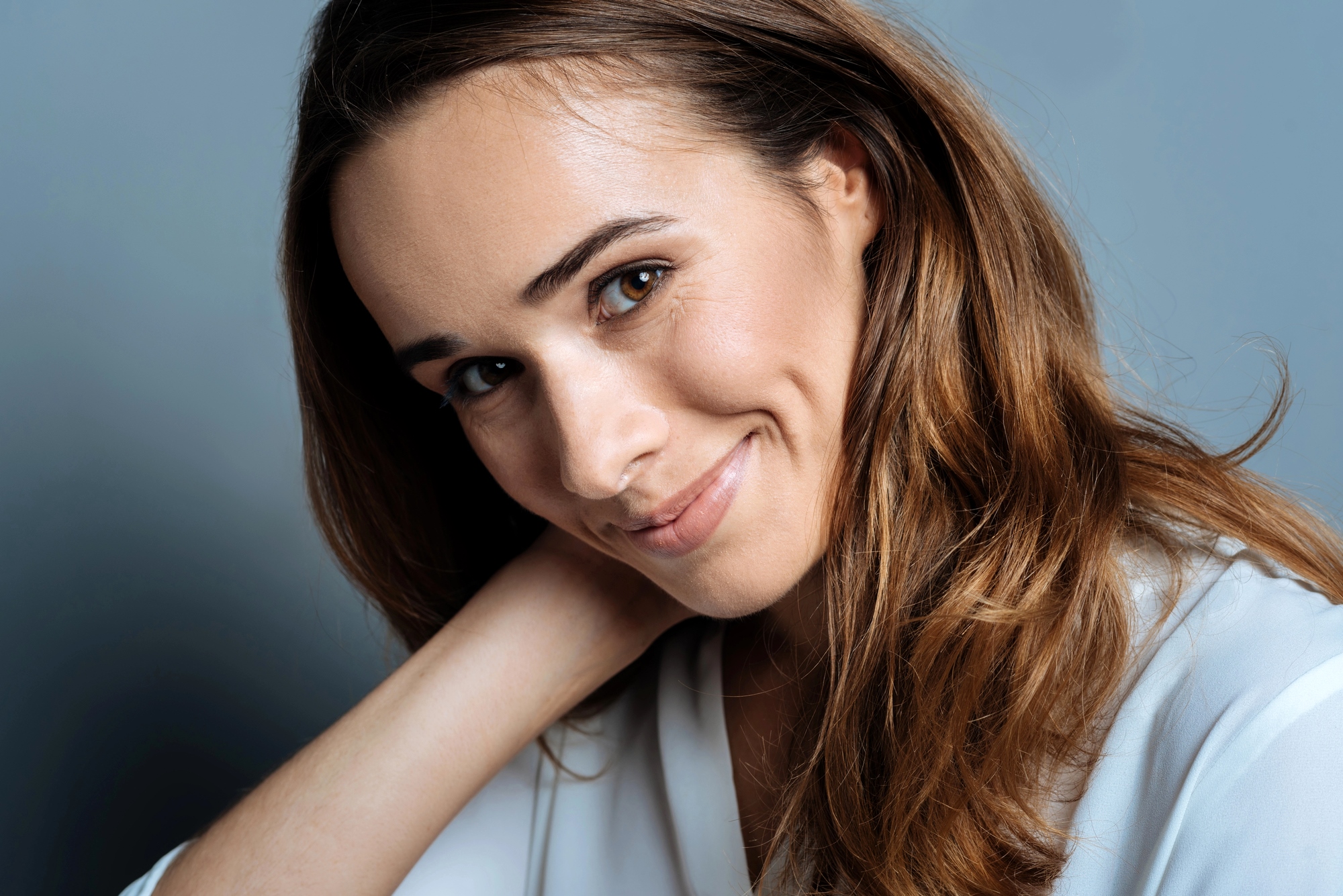 A woman with long brown hair and a light complexion smiles softly while resting her head on her hand. She is wearing a white shirt and has a neutral background.