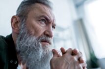 Elderly man with a long gray beard and serious expression, resting his chin on his clasped hands. He is gazing intently into the distance, with a soft, blurred background that suggests an indoor setting. He appears deep in thought.