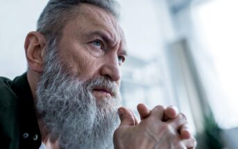 Elderly man with a long gray beard and serious expression, resting his chin on his clasped hands. He is gazing intently into the distance, with a soft, blurred background that suggests an indoor setting. He appears deep in thought.