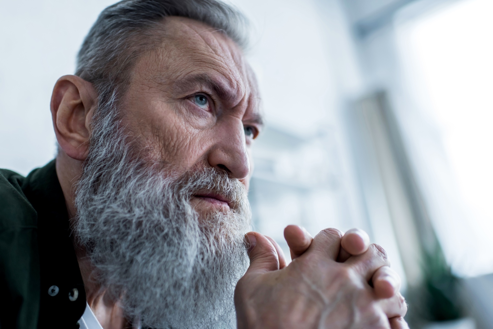 Elderly man with a long gray beard and serious expression, resting his chin on his clasped hands. He is gazing intently into the distance, with a soft, blurred background that suggests an indoor setting. He appears deep in thought.