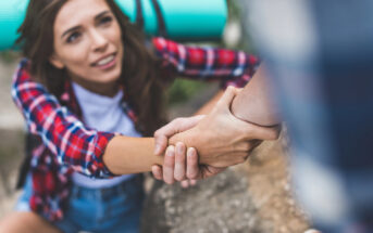 A woman in a plaid shirt and denim shorts is being helped up by another person. She is holding their hand tightly and smiling, with a blue sleeping bag strapped to her back. The background is outdoors, suggesting they might be hiking or camping.