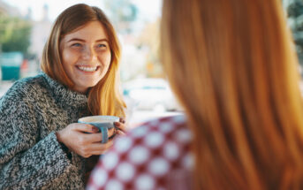 A woman with long red hair, wearing a grey sweater, smiles while holding a cup of coffee. She is facing another person with red hair who is turned away from the camera, wearing a red and white checkered shirt. The background is blurred outdoors.