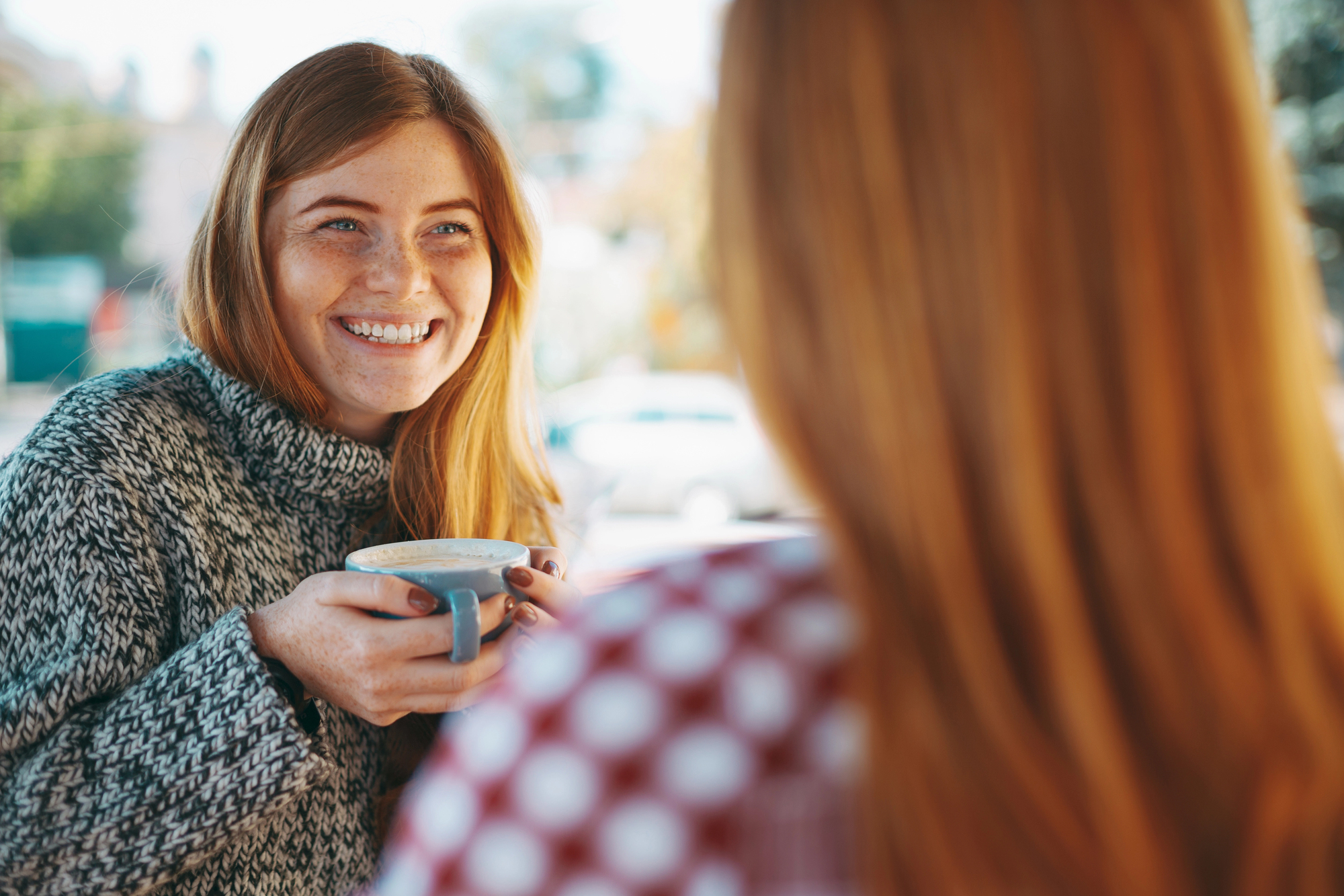 A woman with long red hair, wearing a grey sweater, smiles while holding a cup of coffee. She is facing another person with red hair who is turned away from the camera, wearing a red and white checkered shirt. The background is blurred outdoors.