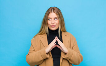 A woman with long blonde hair, wearing a black turtleneck and a tan coat, stands against a bright blue background. She has her hands together in front of her chest and is looking to the side with a playful, mischievous expression on her face.