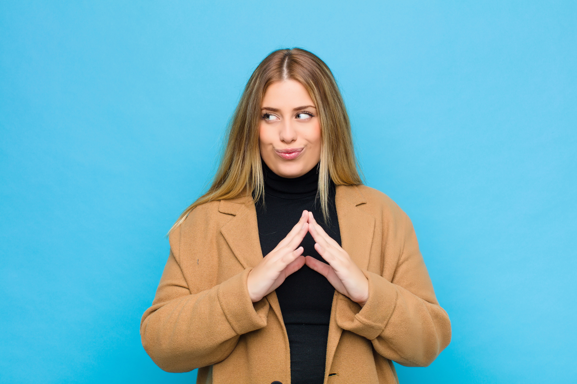 A woman with long blonde hair, wearing a black turtleneck and a tan coat, stands against a bright blue background. She has her hands together in front of her chest and is looking to the side with a playful, mischievous expression on her face.