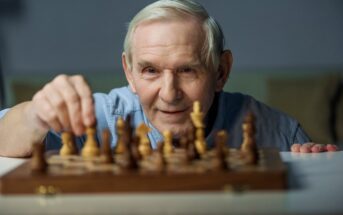 An elderly man with white hair and a blue shirt smiles as he makes a move on a wooden chessboard. He appears to be focused and enjoying the game. The background is blurred, keeping the attention on the man and the chessboard.