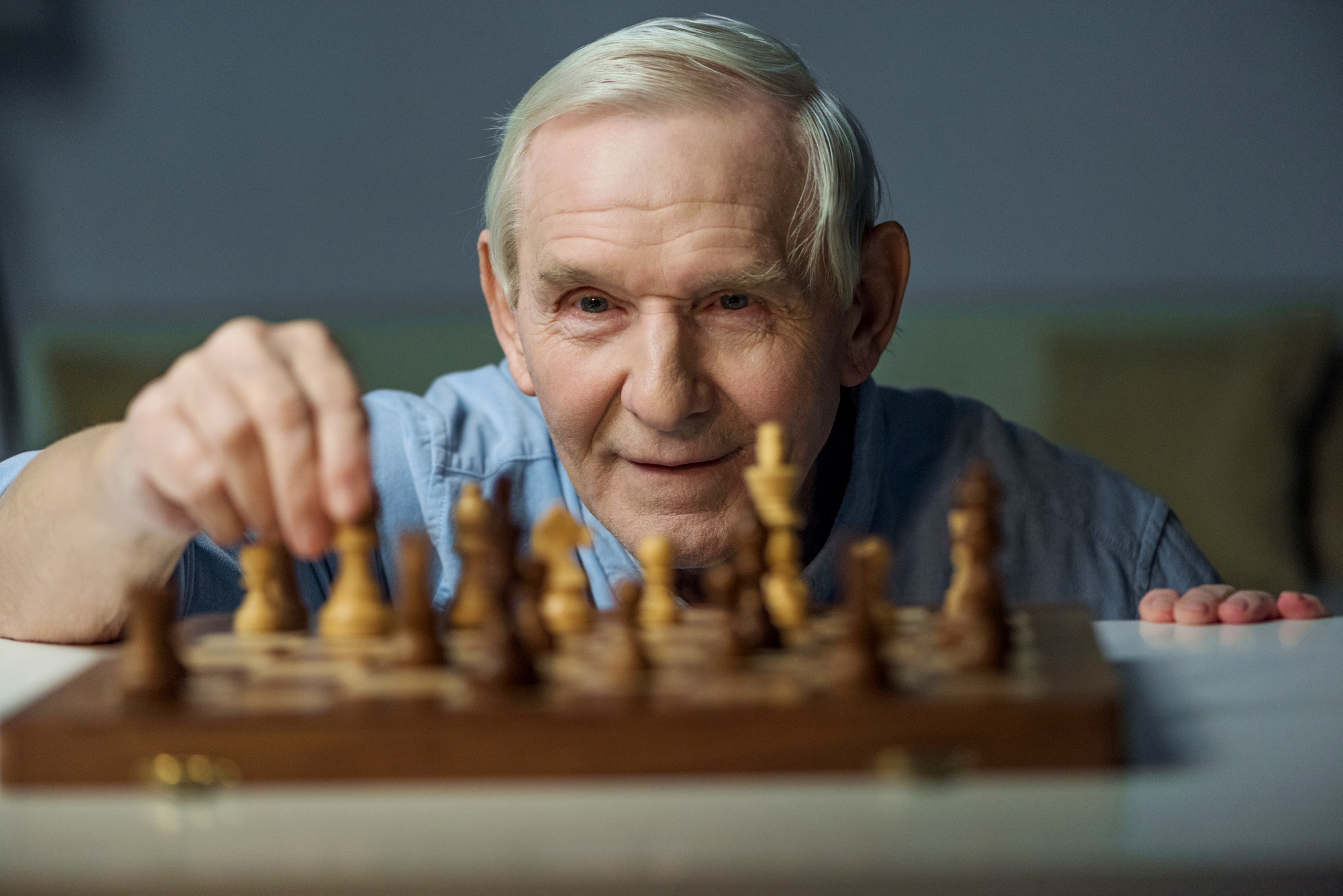 An elderly man with white hair and a blue shirt smiles as he makes a move on a wooden chessboard. He appears to be focused and enjoying the game. The background is blurred, keeping the attention on the man and the chessboard.