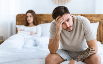 A man sits on the edge of a bed holding his head in his hand, looking distressed. In the background, a woman sits with arms crossed and appears upset. The setting is a bright bedroom with a wooden headboard.