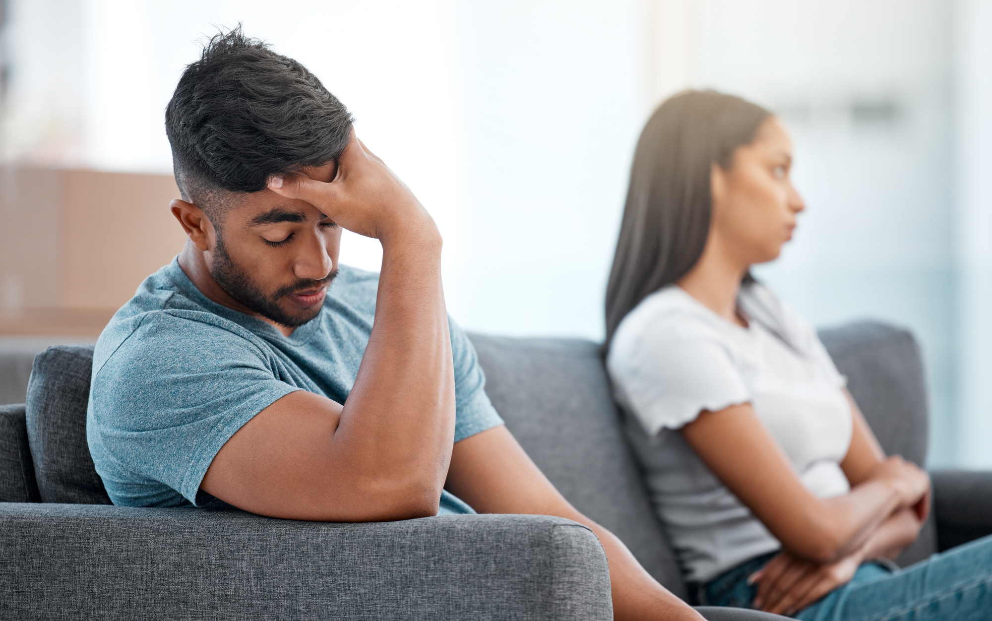 A man and woman sit on a couch facing away from each other. The man, wearing a blue t-shirt, has his hand on his forehead, appearing stressed. The woman, wearing a white t-shirt, looks away with her arms crossed, suggesting they are upset or having an argument.