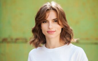 A young woman with wavy, shoulder-length brown hair and a light complexion is standing in front of a blurred green background. She is wearing a white shirt and is smiling gently.