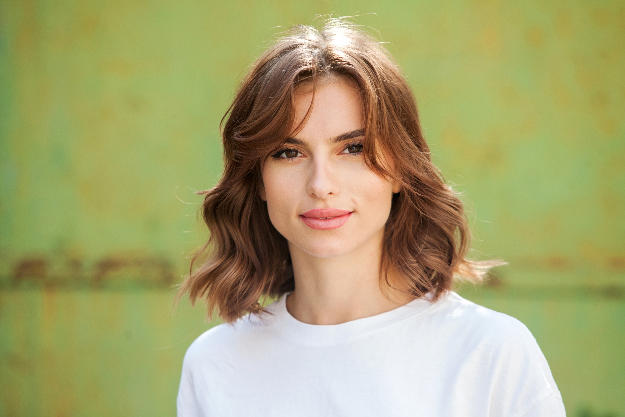 A young woman with wavy, shoulder-length brown hair and a light complexion is standing in front of a blurred green background. She is wearing a white shirt and is smiling gently.