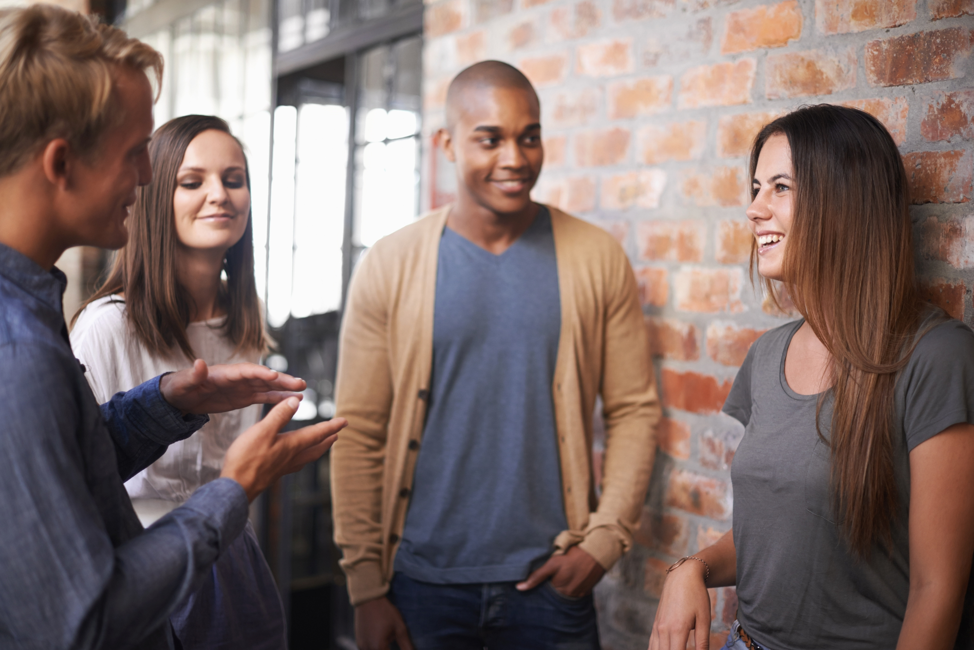 Four people are standing and chatting against a brick wall. One person is talking with hand gestures, while the others are listening and smiling. They appear to be enjoying a conversation in a casual indoor setting.