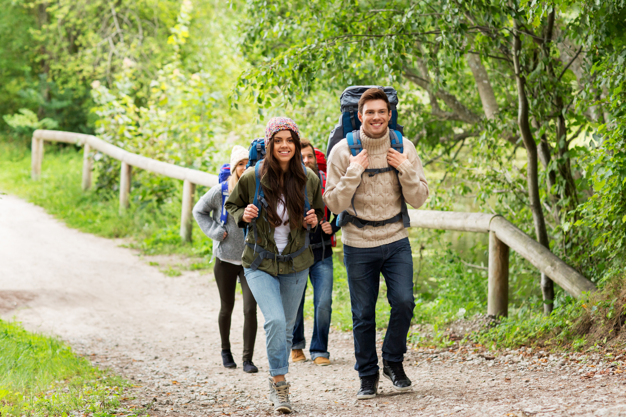 A group of four young adults is hiking along a forest path. They are wearing casual outdoor clothing and backpacks. Trees and greenery surround the path, creating a serene atmosphere. The group appears to be enjoying their time together in nature.