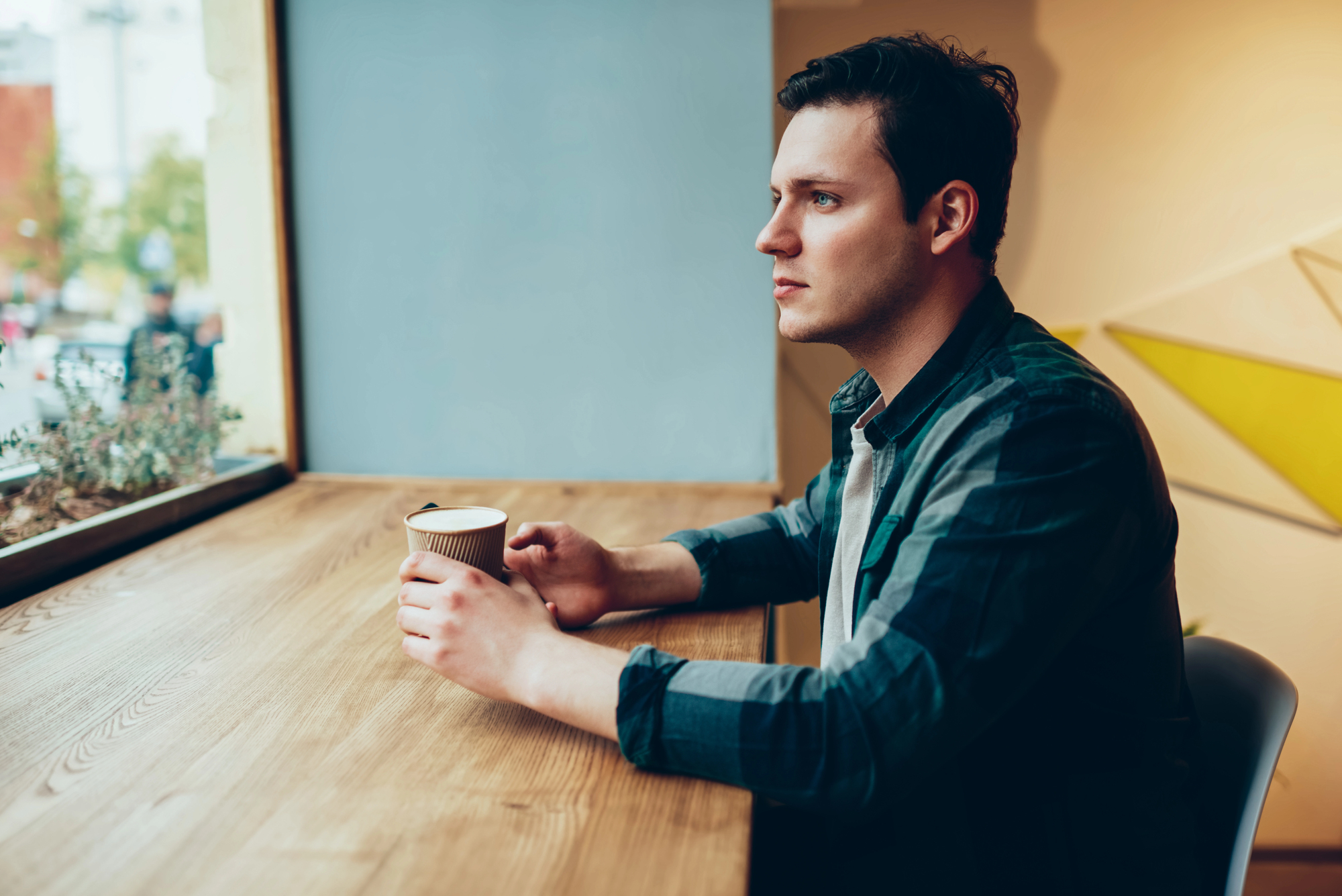 A man with short dark hair sits at a wooden counter by a window, holding a disposable coffee cup with both hands. He looks out the window thoughtfully, wearing a blue and green plaid shirt. The interior has a warm, cozy atmosphere with minimalistic decor.