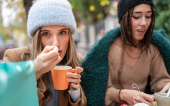 Two people sit at an outdoor cafe. The person on the left, wearing a light blue beanie and brown coat, eats from an orange mug, looking at the camera. The person on the right, in a black beanie and wrapped in a green blanket, looks down at the table.