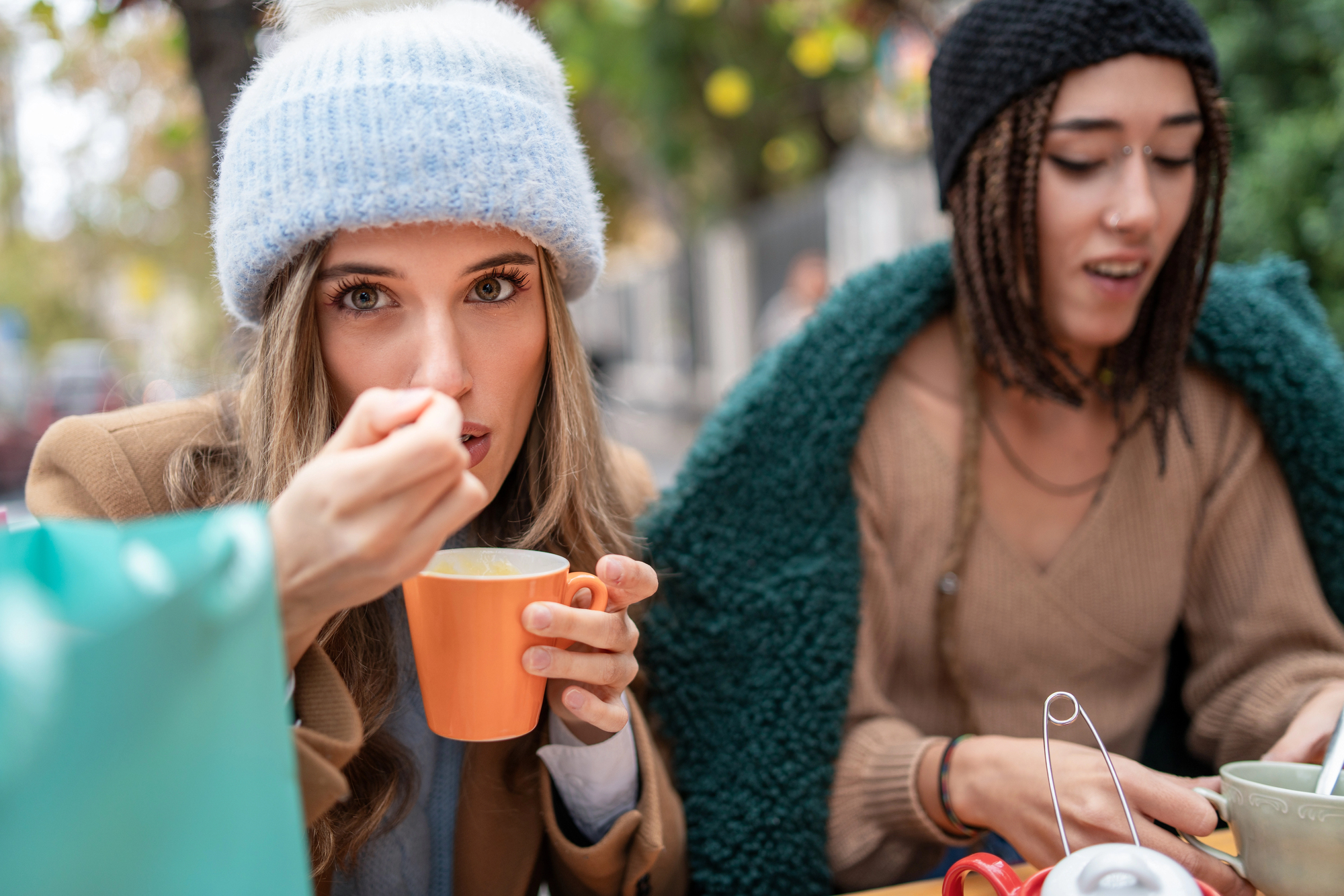 Two people sit at an outdoor cafe. The person on the left, wearing a light blue beanie and brown coat, eats from an orange mug, looking at the camera. The person on the right, in a black beanie and wrapped in a green blanket, looks down at the table.