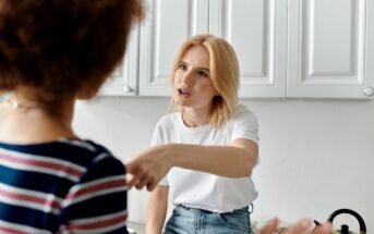 A woman with blonde hair dressed in a white T-shirt and jeans is gesturing and appears to be expressing frustration or arguing with another person. The second person, who has curly hair and is wearing a striped shirt, has their back to the camera. They are in a kitchen.