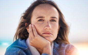 A woman with long brown hair rests her chin on her hand, looking pensive. She is wearing a blue denim jacket and has a thoughtful expression. The background is blurred and bright, suggesting an outdoor setting with sunlight.