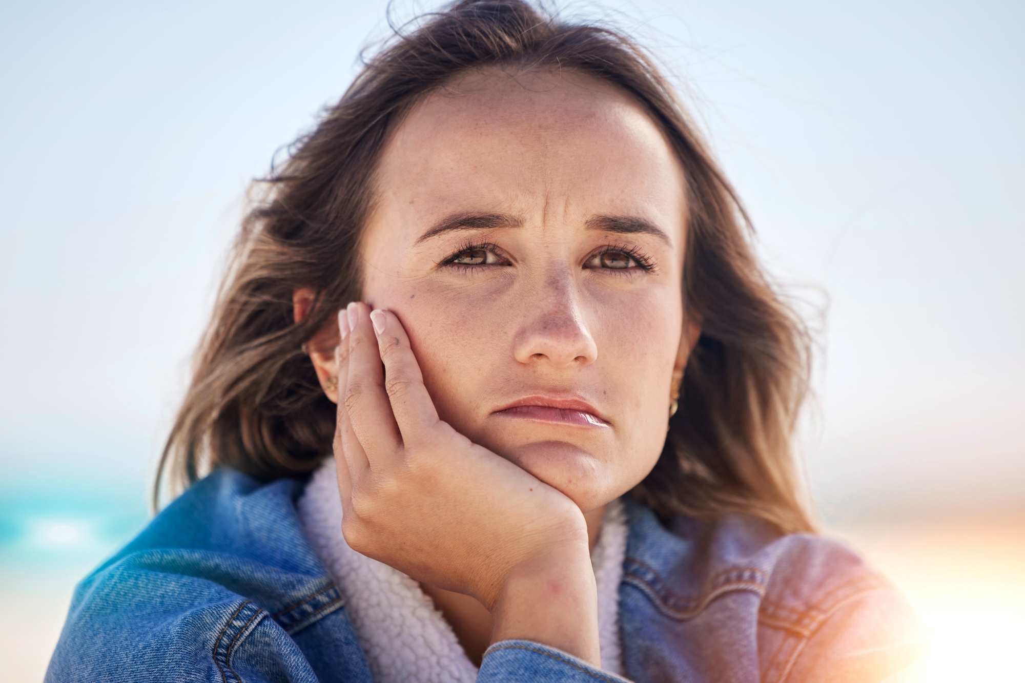 A woman with long brown hair rests her chin on her hand, looking pensive. She is wearing a blue denim jacket and has a thoughtful expression. The background is blurred and bright, suggesting an outdoor setting with sunlight.