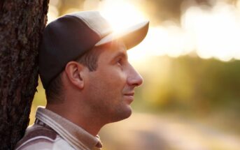 A man wearing a baseball cap leans against a tree with sunlight streaming through the background. He is looking off to the side with a peaceful expression. The background is softly blurred, creating a warm, serene atmosphere.