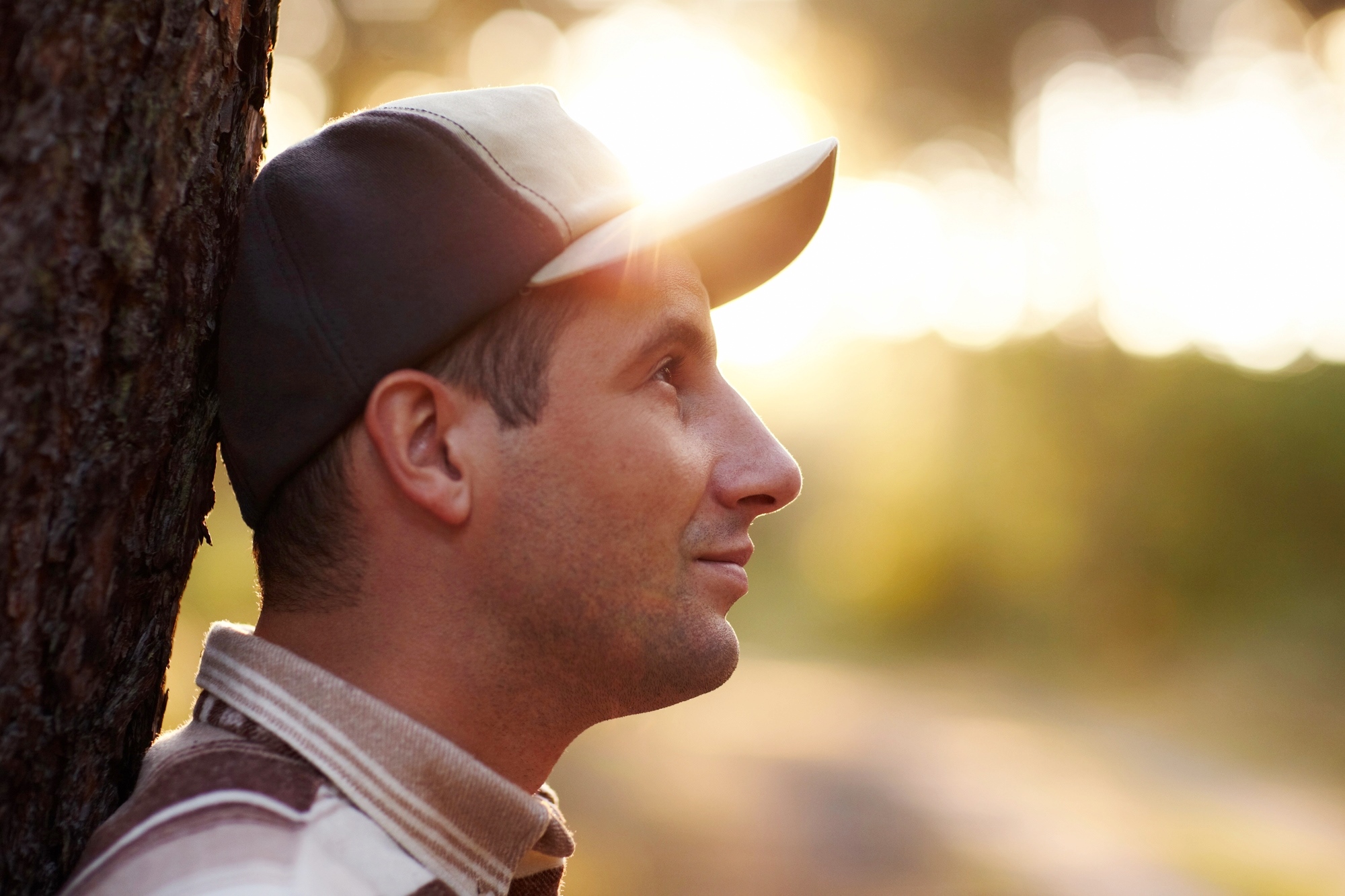 A man wearing a baseball cap leans against a tree with sunlight streaming through the background. He is looking off to the side with a peaceful expression. The background is softly blurred, creating a warm, serene atmosphere.