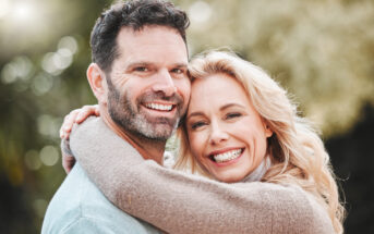 A man and a woman are outdoors, smiling and embracing. The woman has blonde hair and is wearing a light brown sweater, while the man has dark hair, a beard, and is wearing a light blue sweater. The background is blurred green foliage.