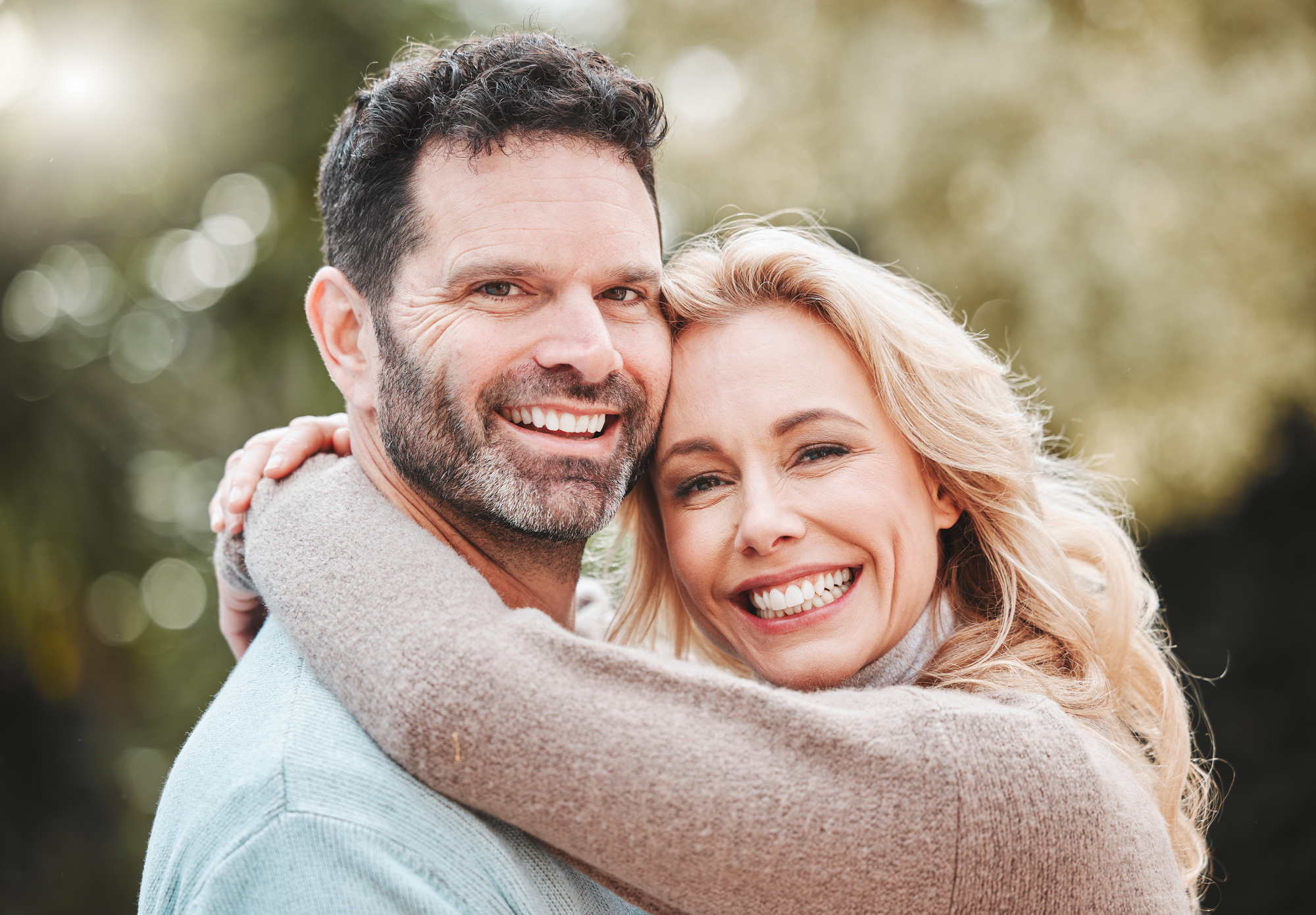 A man and a woman are outdoors, smiling and embracing. The woman has blonde hair and is wearing a light brown sweater, while the man has dark hair, a beard, and is wearing a light blue sweater. The background is blurred green foliage.