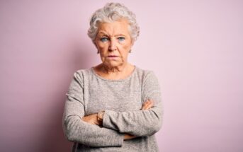 An elderly woman with short, curly gray hair stands against a pale pink background with her arms crossed. She wears a light gray sweater and has a serious, determined expression on her face.