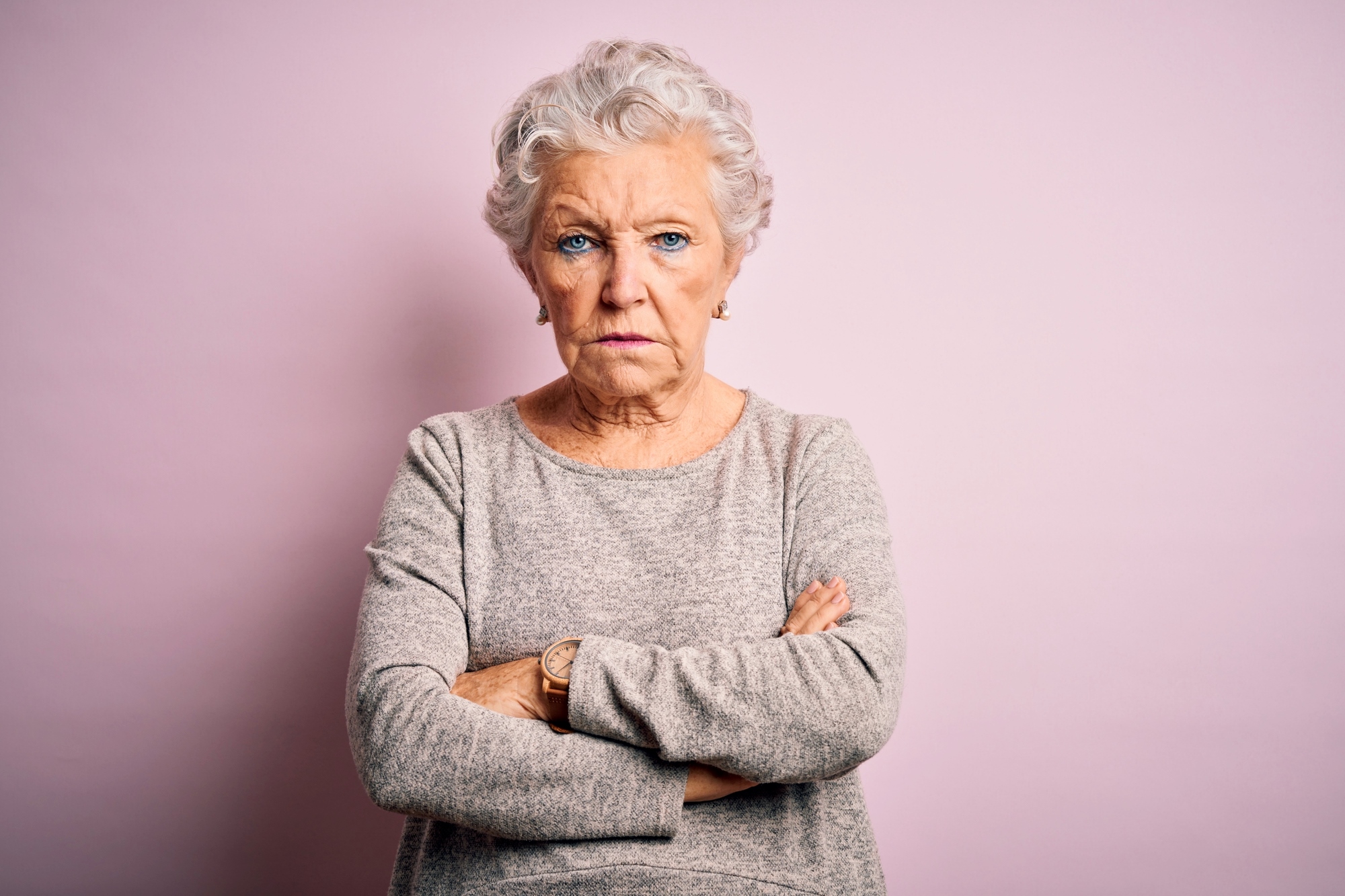 An elderly woman with short, curly gray hair stands against a pale pink background with her arms crossed. She wears a light gray sweater and has a serious, determined expression on her face.