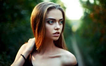 A young woman with long, straight hair stands outdoors, looking to her left with a serious expression. She is wearing an off-the-shoulder black top and has her hand on her neck. The background is a blurred natural setting of greenery.