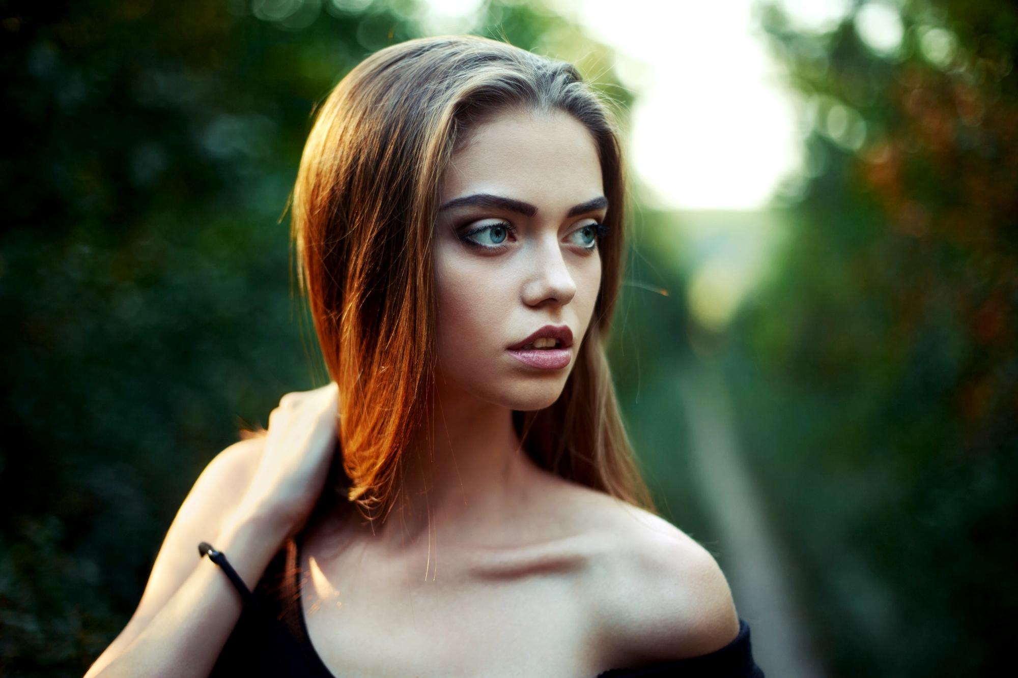 A young woman with long, straight hair stands outdoors, looking to her left with a serious expression. She is wearing an off-the-shoulder black top and has her hand on her neck. The background is a blurred natural setting of greenery.